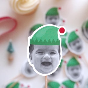 Puede incluir: Una colección de recortes de caras de bebés sonrientes con sombreros de elfo verdes con un pompón rojo. Los recortes están adheridos a palitos de madera, probablemente para usar como adornos para cupcakes o decoraciones de fiestas. El fondo está borroso.