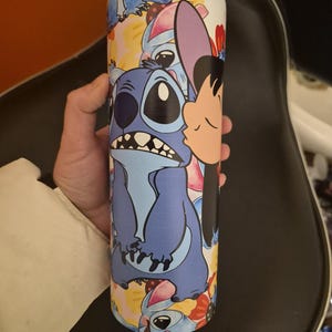 Puede incluir: Un vaso de acero inoxidable con un dise&ntilde;o de dibujos animados de Stitch de Lilo & Stitch. El vaso tiene una tapa negra y est&aacute; cubierto de im&aacute;genes de Stitch en varias poses, con un fondo blanco.