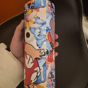 Puede incluir: Un vaso cil&iacute;ndrico con un colorido dise&ntilde;o del personaje Stitch de la pel&iacute;cula animada Lilo & Stitch. El dise&ntilde;o incluye a Stitch en varias poses, sobre un fondo de flores y otros elementos. El vaso es sostenido por una persona.