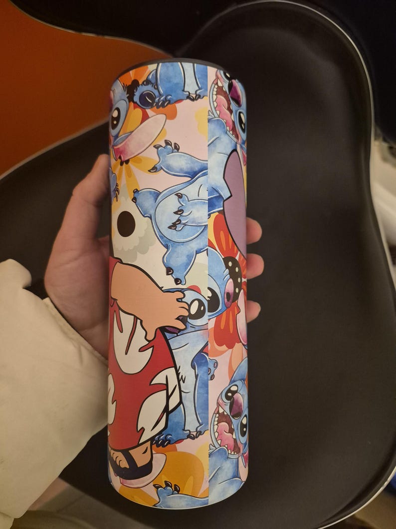 Puede incluir: Un vaso cil&iacute;ndrico con un colorido dise&ntilde;o del personaje Stitch de la pel&iacute;cula animada Lilo & Stitch. El dise&ntilde;o incluye a Stitch en varias poses, sobre un fondo de flores y otros elementos. El vaso es sostenido por una persona.