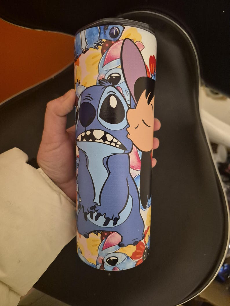 Puede incluir: Un vaso de acero inoxidable con un dise&ntilde;o de dibujos animados de Stitch de Lilo & Stitch. El vaso tiene una tapa negra y est&aacute; cubierto de im&aacute;genes de Stitch en varias poses, con un fondo blanco.