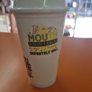 May include: A white plastic travel tumbler with a matching white lid. The cup features a colorful graphic print with the text: If my MOUTH DOESN'T SAY IT my face DEFINITELY WILL. The design includes yellow, purple, and black lettering.