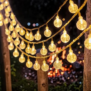 Puede incluir: Guirnalda de luces con pequeñas bombillas redondas y transparentes, que emiten un brillo amarillo cálido. Las luces están ensartadas en un cable dorado, creando un ambiente festivo y acogedor. El fondo es oscuro, con destellos de fuego.