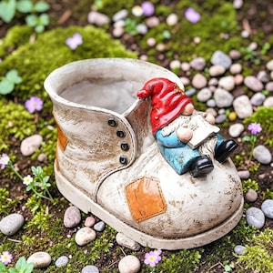 Puede incluir: Una maceta en forma de bota desgastada, de color blanquecino, con parches marrones y ojales negros. Una figura de gnomo con gorro rojo y chaqueta azul descansa sobre la bota, sosteniendo un libro. Rodeada de musgo, piedras y flores.