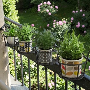 Puede incluir: Cuatro plantas en macetas en soportes decorativos fijados a una barandilla de metal negro. Las macetas tienen varios patrones y colores, incluyendo blanco, amarillo y gris. El fondo presenta un jardín con flores rosas y follaje verde.