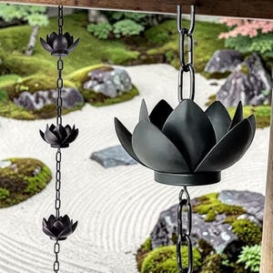 Op de afbeelding: Zwarte metalen lotusbloem regenkettingen die aan een houten balk hangen. De kettingen zijn gemaakt van metalen schakels en de lotusbloemen zijn gedetailleerd. De achtergrond toont een Japanse tuin met rotsen en groen.