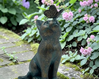 Puede incluir: Estatua de gato de bronce con una mariposa en la cabeza. El gato está sentado y mirando hacia arriba. La estatua tiene un aspecto envejecido, lo que sugiere su uso en exteriores. El fondo muestra un jardín con flores.