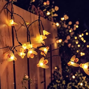 May include: A string of bee-shaped fairy lights, with a warm yellow and orange glow. The lights are strung against a wooden fence, with a dark background of foliage and other lights. The lights are ideal for outdoor decoration.