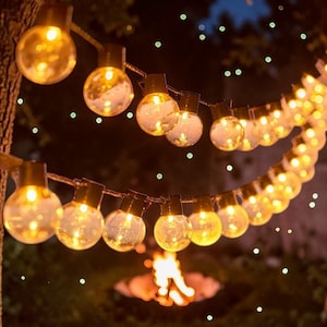 May include: A string of warm, glowing light bulbs hangs against a dark backdrop. The round bulbs are illuminated, creating a cosy atmosphere. The lights are suspended from a tree branch, with a fire pit visible in the background.