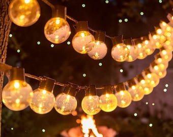 May include: String lights with warm, glowing bulbs are strung across a dark background. The round bulbs are lit, creating a cozy ambiance. The lights are hanging from a tree branch, with a fire pit visible in the background.
