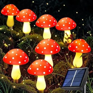 Puede incluir: Un conjunto de luces solares de jardín con forma de hongo rojo con lunares blancos. Las luces tienen tallos amarillos y están dispuestas entre vegetación. También se ve un panel solar.