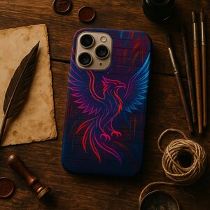 May include: A phone case with a vibrant phoenix design in red, pink, and blue hues. The case has a dark blue background with a textured appearance. The image is set on a wooden surface with artistic tools and stationery.