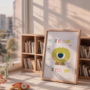 May include: A framed wall art print featuring a fluffy yellow monster with one large blue eye. The text reads IT IS OKAY TO FEEL SHY. The design includes small illustrations of clouds, stars, and rainbows on a white background within a light wood frame.
