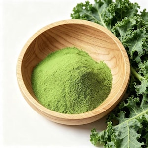 Puede incluir: Un montón de polvo de col rizada verde vibrante contenido en un cuenco de madera lisa de color claro, colocado junto a un racimo de hojas de col rizada frescas y rizadas con bordes texturizados, sobre un fondo blanco limpio.
