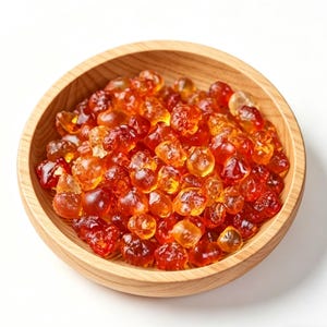 Peut inclure: Un bol en bois rempli de morceaux translucides et de forme irrégulière de couleur ambre. Le bol est en bois clair et repose sur une surface blanche. Le contenu a un aspect brillant, avec des variations de couleur du jaune clair au rouge foncé.
