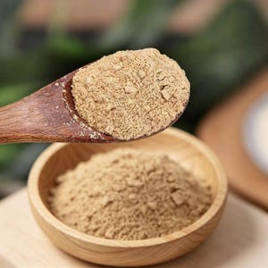 May include: A wooden spoon holds a mound of fine, light brown powder, positioned above a small, round wooden bowl filled with the same textured, beige-coloured substance. The wooden grain of the spoon and bowl is visible in this close-up view.