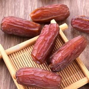 Può includere: Diverse datteri marroni essiccati con la buccia rugosa e strutturata sono disposti su un vassoio di bambù chiaro. I frutti di forma ovale presentano una profonda tonalità bruno-rossastra e una finitura lucida, che ne evidenzia l'aspetto naturale.