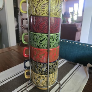 Puede incluir: Un juego de seis tazas de cerámica apiladas en un soporte vertical de metal cromado. Cada taza presenta un patrón de cachemira en espiral de color marrón oscuro sobre un fondo de color sólido diferente, incluyendo verde lima, rojo oscuro, verde claro, naranja, azul marino y amarillo.