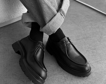 May include: A pair of black leather shoes with a moc-toe design, thin laces, and a thick, lugged rubber sole. The shoes are worn with black socks and cuffed grey trousers, showcasing a structured silhouette with visible stitching along the vamp.