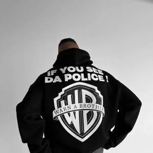 Könnte beinhalten: Schwarzer Kapuzenpullover mit weißem Text und einem Schildlogo. Der Text auf der Rückseite lautet "IF YOU SEE DA POLICE! WARN A BROTHER" über und unter einem stilisierten "WB"-Logo.