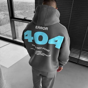 May include: A grey hoodie and matching sweatpants. The back of the hoodie displays the text "ERROR 404" in light blue. Underneath, the words "HUMAN NOT FOUND" and "SORRY! YOU ARE LOOKING FOR IS NOT EXIST" are printed.