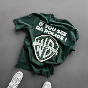 Könnte beinhalten: Ein dunkelgrünes kurzärmeliges T-Shirt mit weißem Text, der IF YOU SEE DA POLICE! lautet, über einem weißen grafischen Logo in Schildform mit dem Text WARN A BROTHER und den Buchstaben WD darin. Das T-Shirt wird mit weißen Turnschuhen kombiniert.