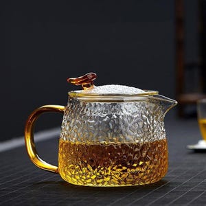 Puede incluir: Una tetera de vidrio transparente con una superficie texturizada, llena de líquido ámbar. La tetera tiene un asa dorada y una tapa con un pequeño asa decorativa. El diseño es elegante y adecuado para servir té.