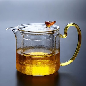 May include: A clear glass teapot with a gold-colored handle and lid. The teapot is filled with a golden-colored liquid. The lid has a small, amber-colored knob. The teapot has a textured surface and a spout for pouring.