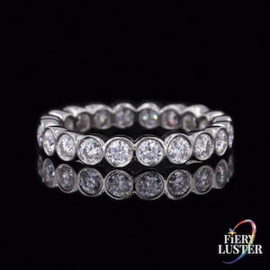 Puede incluir: Un anillo de eternidad en tono plateado que presenta una fila continua de piedras redondas y transparentes engastadas en bisel. El anillo está colocado horizontalmente, mostrando una superficie reflectante y un diseño circular. El texto Fiery Luster es visible.