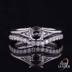 May include: A silver engagement ring set with a round center setting and a curved band with small, sparkling stones. The ring is reflected on a black surface, showcasing the intricate design and craftsmanship. The ring is from Fiery Luster.