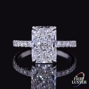 May include: A radiant-cut diamond ring featuring a large, rectangular centre stone held by four prongs, set on a thin, polished metal band encrusted with a row of small, round, brilliant-cut diamonds that extend along the sides of the ring.