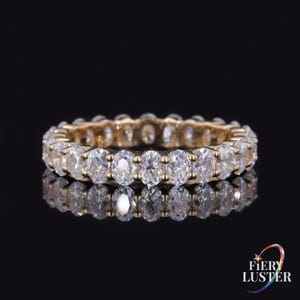 Könnte beinhalten: Ein Eternity-Ring mit einer durchgehenden Reihe von oval geschliffenen, klaren Steinen in einer goldfarbenen Metallschiene. Der Ring ist vor einem schwarzen Hintergrund abgebildet, mit einer Spiegelung darunter und dem Text Fiery Luster in der Ecke.