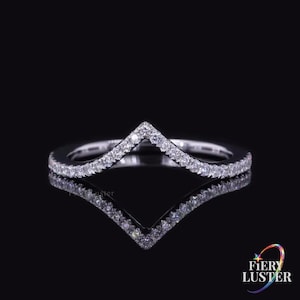 Peut inclure: Une bague en forme de chevron argentée ornée d'une rangée continue de petites pierres claires et scintillantes serties le long de toute la bande en V. La bague est présentée sur un fond noir avec un reflet net visible sous l'objet.