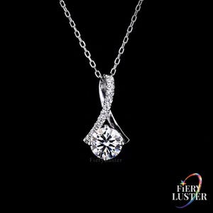 May include: A silver-toned necklace featuring a pendant with a large, round, clear faceted stone at the center. The pendant has a twisted, ribbon-like design accented with small, clear stones along one side. The text Fiery Luster is visible.