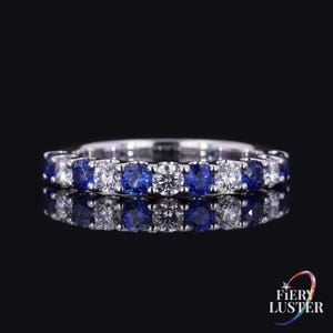 May include: A silver-toned eternity band ring featuring an alternating pattern of round-cut blue sapphires and clear white diamonds set in a shared prong setting. The ring is displayed against a black background with a clear reflection below it.