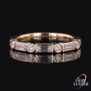 Op de afbeelding: Een gouden ring met een afwisselend patroon van heldere baguette-geslepen stenen en ronde briljant-geslepen stenen in een zetting. De ring heeft een gepolijste metalen afwerking en reflecteert het licht op het donkere oppervlak.