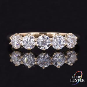 Puede incluir: Un anillo de oro con cinco piedras redondas transparentes dispuestas en fila en la parte superior. Las piedras están montadas en engaste de garras, creando una superficie reflectante. El texto Fiery Luster es visible en la esquina inferior.