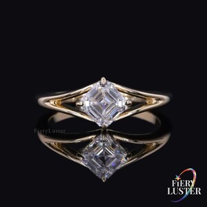 May include: A gold ring featuring a square-cut, clear gemstone. The ring's band splits into a double-band design on either side of the gemstone. The ring is set against a black background, with a reflection of the ring visible below.