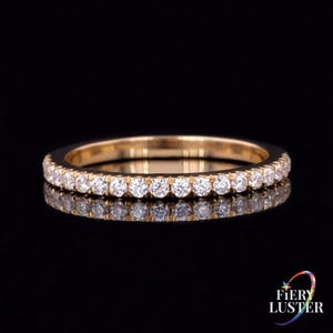 Può includere: Un sottile anello a fascia in oro con una fila continua di piccole pietre rotonde trasparenti sfaccettate incastonate sulla superficie esterna. L'anello ha una finitura metallica lucida e riflette la luce, creando un aspetto brillante su sfondo scuro.