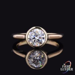 May include: A gold ring with a large, round, clear gemstone set in a bezel setting. The ring is displayed on a reflective black surface, creating a mirrored effect. The ring's band is smooth and simple.