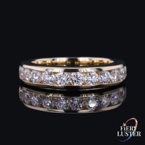 Puede incluir: Un anillo de banda de oro con un engaste de canal que presenta una fila continua de piedras redondas transparentes de talla brillante. El anillo tiene un acabado metálico pulido y se muestra sobre un fondo oscuro con un reflejo visible debajo.