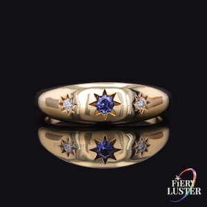 Op de afbeelding: Een gouden ring met een gepolijste afwerking, met drie stervormige zettingen. De middelste zetting bevat een blauwe edelsteen, geflankeerd door twee zettingen met heldere edelstenen. De ring wordt gepresenteerd op een reflecterend oppervlak.