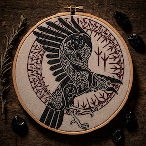 Puede incluir: Arte de cuervo bordado en un aro de madera. El cuervo es negro con intrincados detalles de nudos, sobre un fondo de tela beige. Un diseño circular con detalles en rojo rodea al cuervo. El aro de madera es de color madera natural.