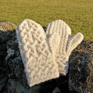 Puede incluir: Un par de guantes de punto beige con un patrón de trenza. Los guantes están sobre una roca con un campo verde y ovejas en el fondo.