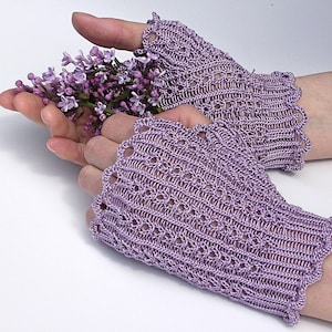 Könnte beinhalten: Ein Paar lavendelfarbene, gehäkelte Fingerlose Handschuhe mit einem Spitzenmuster. Die Handschuhe werden von einer Person getragen, die einen Strauß lilafarbener Fliederblüten hält.