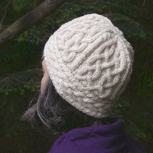 May include: A white knitted hat with a cable knit pattern. The hat is worn by a person with gray hair and a purple jacket.