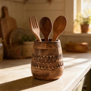 May include: A wooden utensil holder with a carved leaf design, holding a wooden fork and two spoons. The holder is brown and sits on a kitchen countertop. The kitchen has a rustic, farmhouse style.
