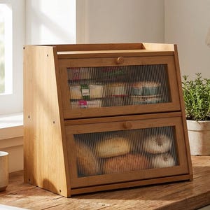 May include: A two-tiered wooden bread box with glass doors. The box is made of light-colored wood and has a simple, rectangular design. The glass doors have a textured, ribbed pattern. The top compartment contains packaged goods, and the bottom compartment holds bread and bagels.