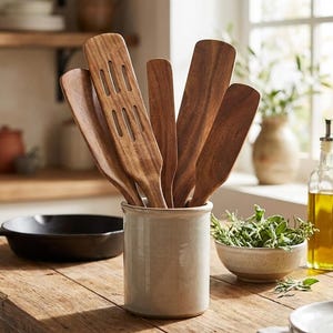 May include: A collection of wooden kitchen utensils, including spatulas and slotted spoons, displayed in a ceramic utensil holder. The utensils are various shades of brown and are arranged on a wooden table. A cast iron skillet and a bowl of herbs are also visible.