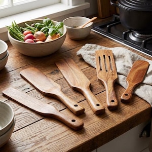 May include: Wooden kitchen utensils, including spatulas and a slotted turner, are arranged on a wooden countertop. A bowl of fresh vegetables and a cast iron pot are also visible. The scene is bathed in natural light, creating a warm, inviting atmosphere.